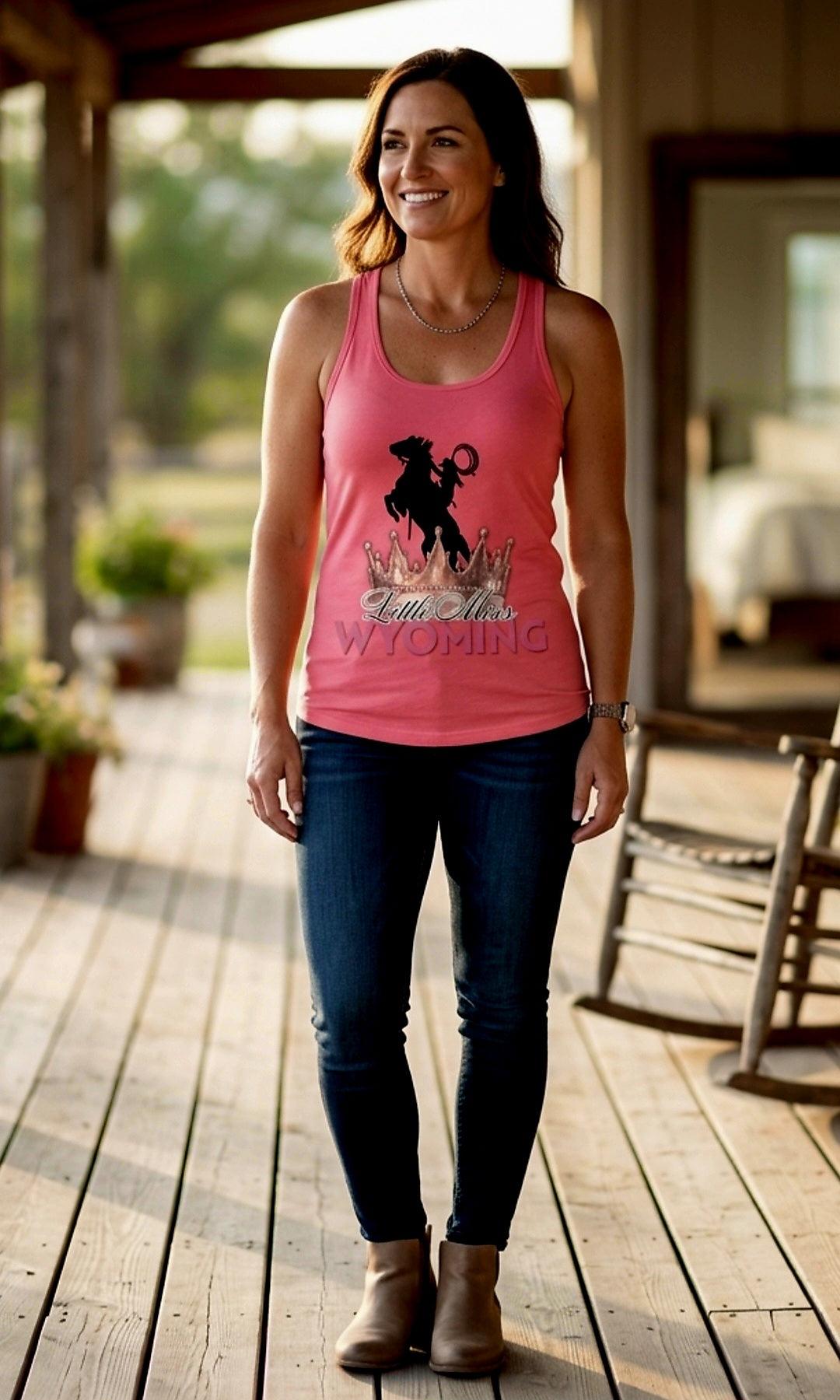 Woman wearing pink 'Little Miss Wyoming' tank top with rodeo rider graphic, standing on rustic porch
