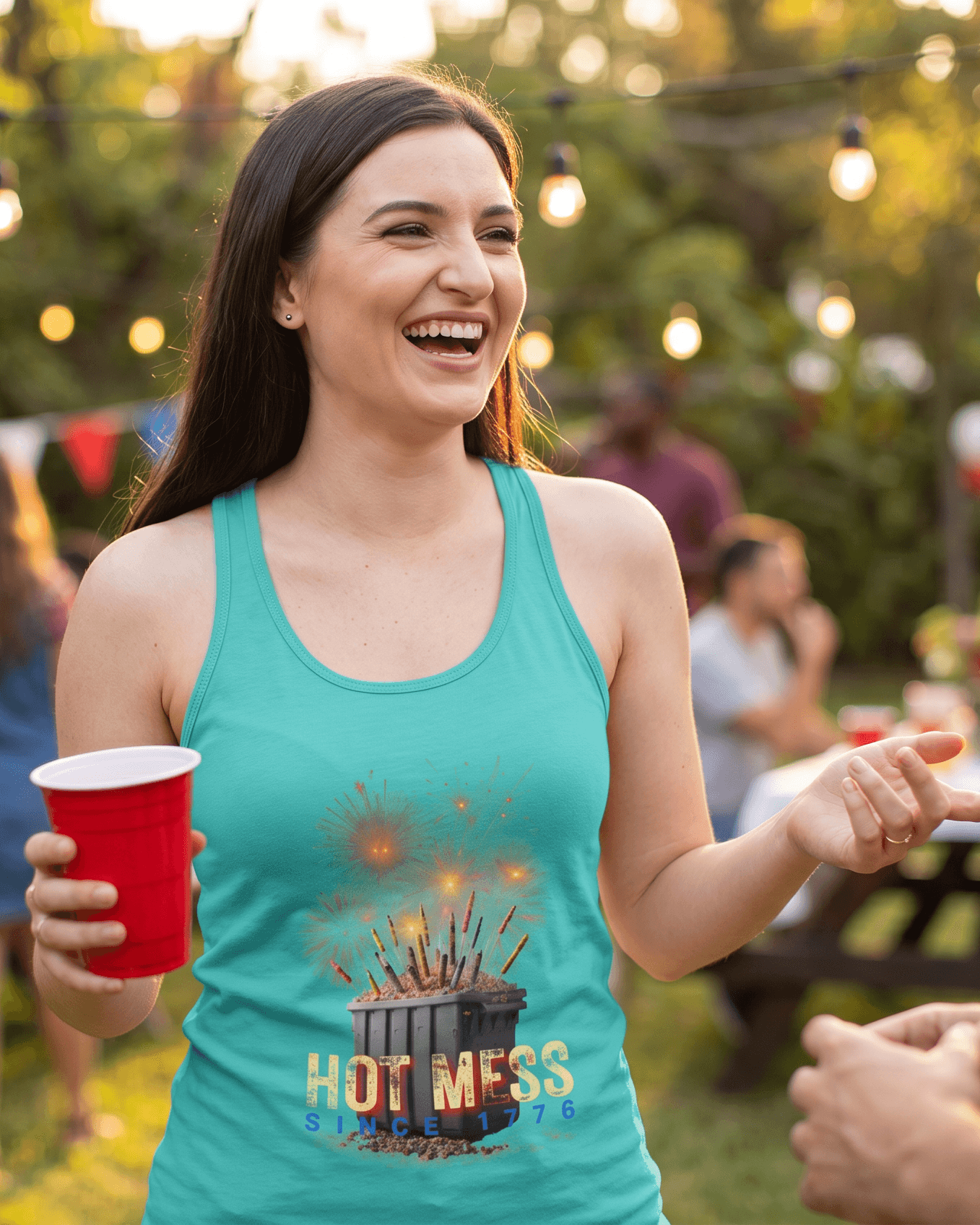 Woman in teal tank top with 'Hot Mess' graphic holding a red cup, standing outdoors at a social gathering.