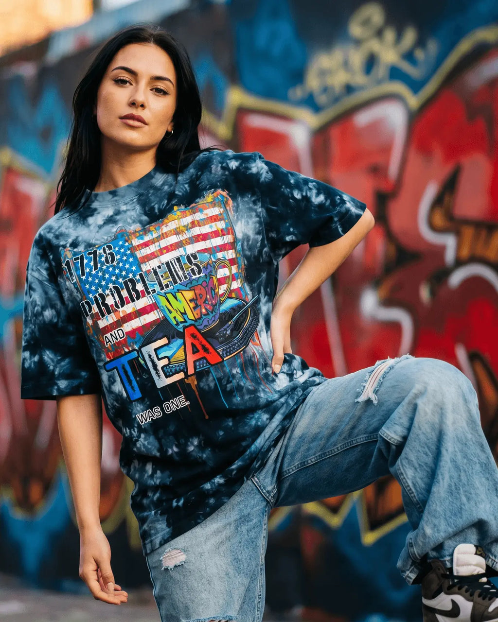 Person wearing a tie-dye shirt with text in front of a graffiti-covered wall