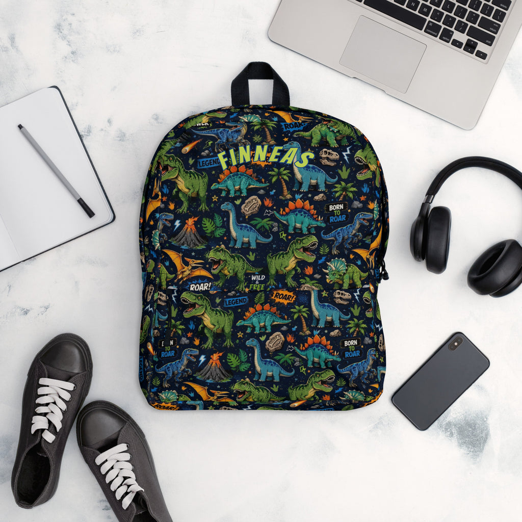 A backpack with a colorful dinosaur pattern is shown in a styled flat-lay arrangement on a white marble surface, surrounded by a laptop, notebook, headphones, phone, and shoes.