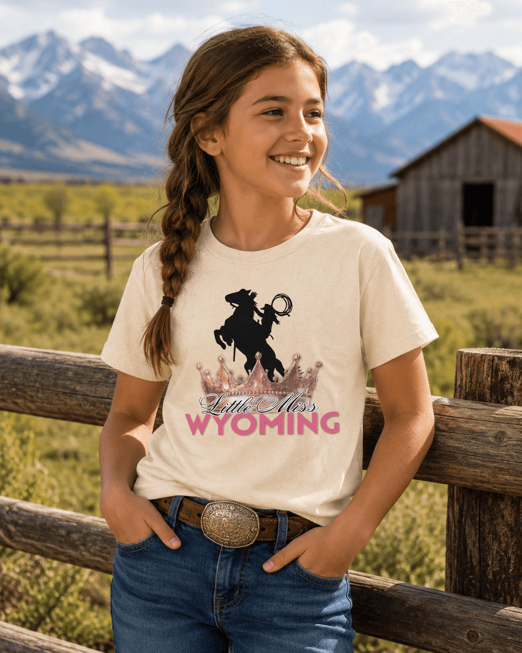 Girl wearing 'Little Miss Wyoming' graphic tee with rodeo rider and crown, set against mountain ranch backdrop