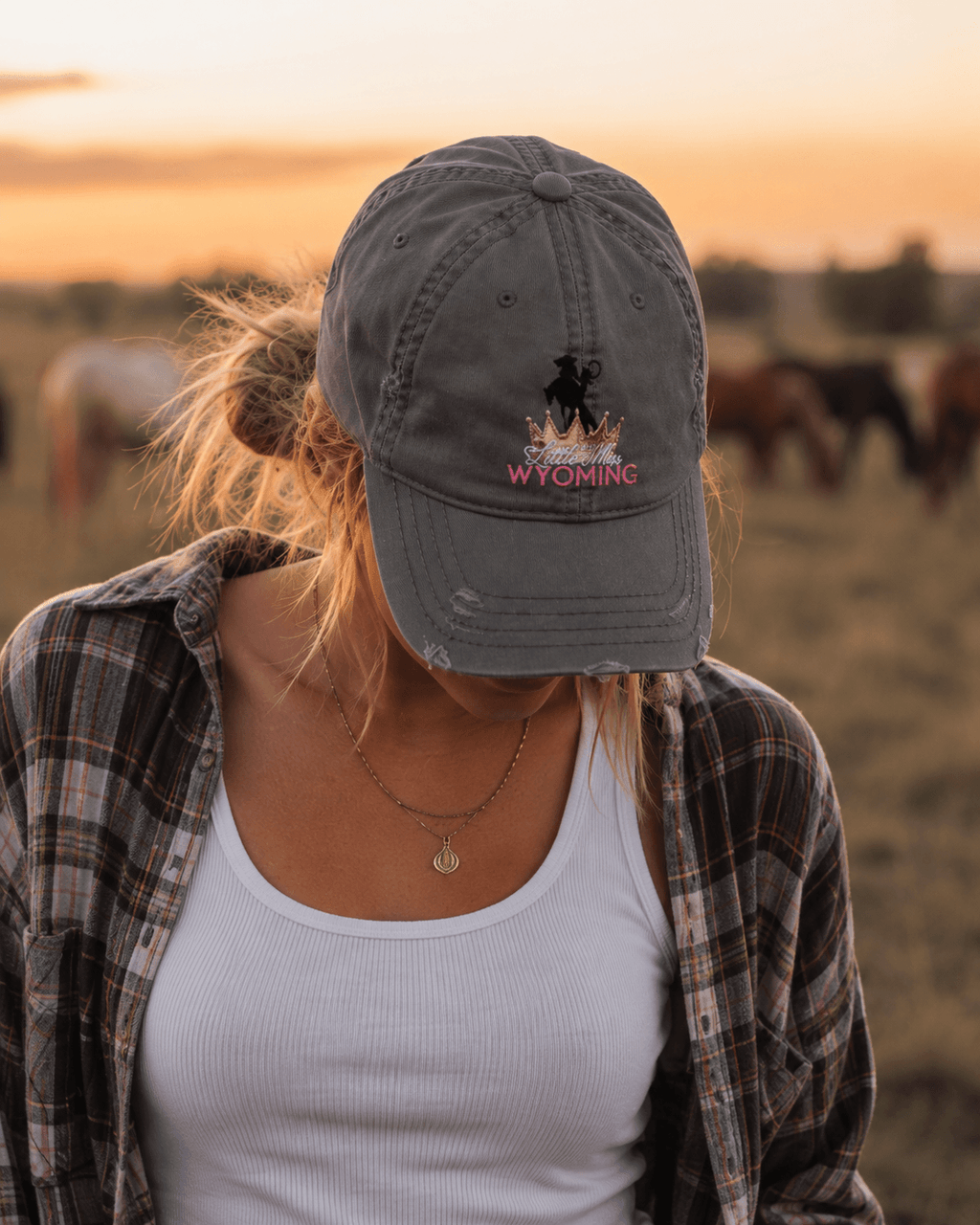 Woman wearing 'Little Miss Wyoming' embroidered hat at sunset with horses, casual plaid shirt outfit