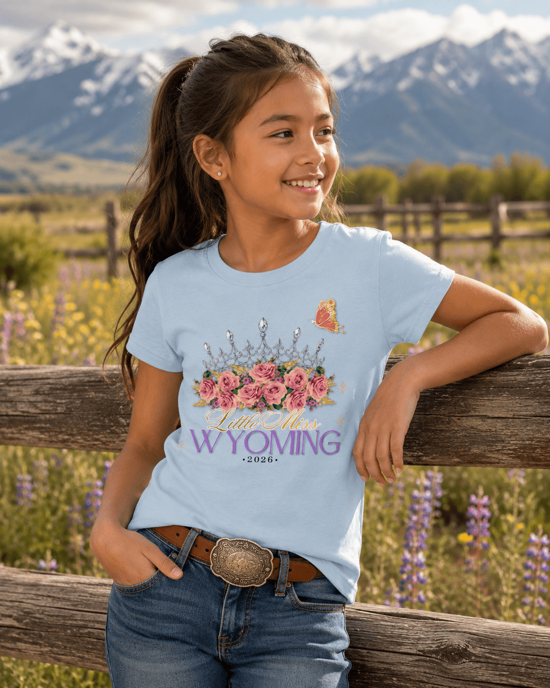 Girl wearing 'Little Miss Wyoming 2026' graphic tee with floral crown, butterfly, and mountain backdrop