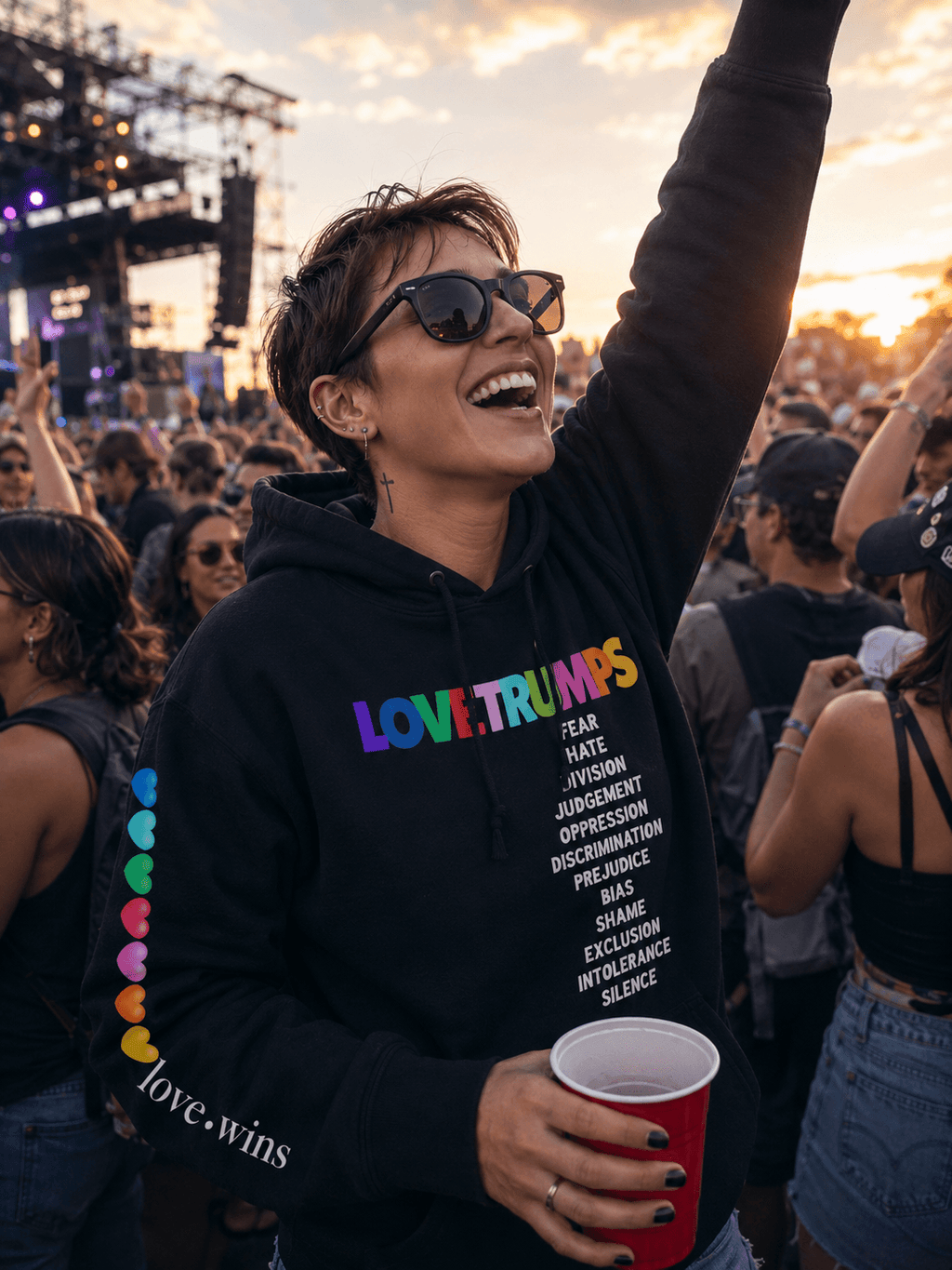 Woman at concert wearing black hoodie with 'LOVE.TRUMPS' rainbow text, colorful hearts, and advocacy message, holding red cup