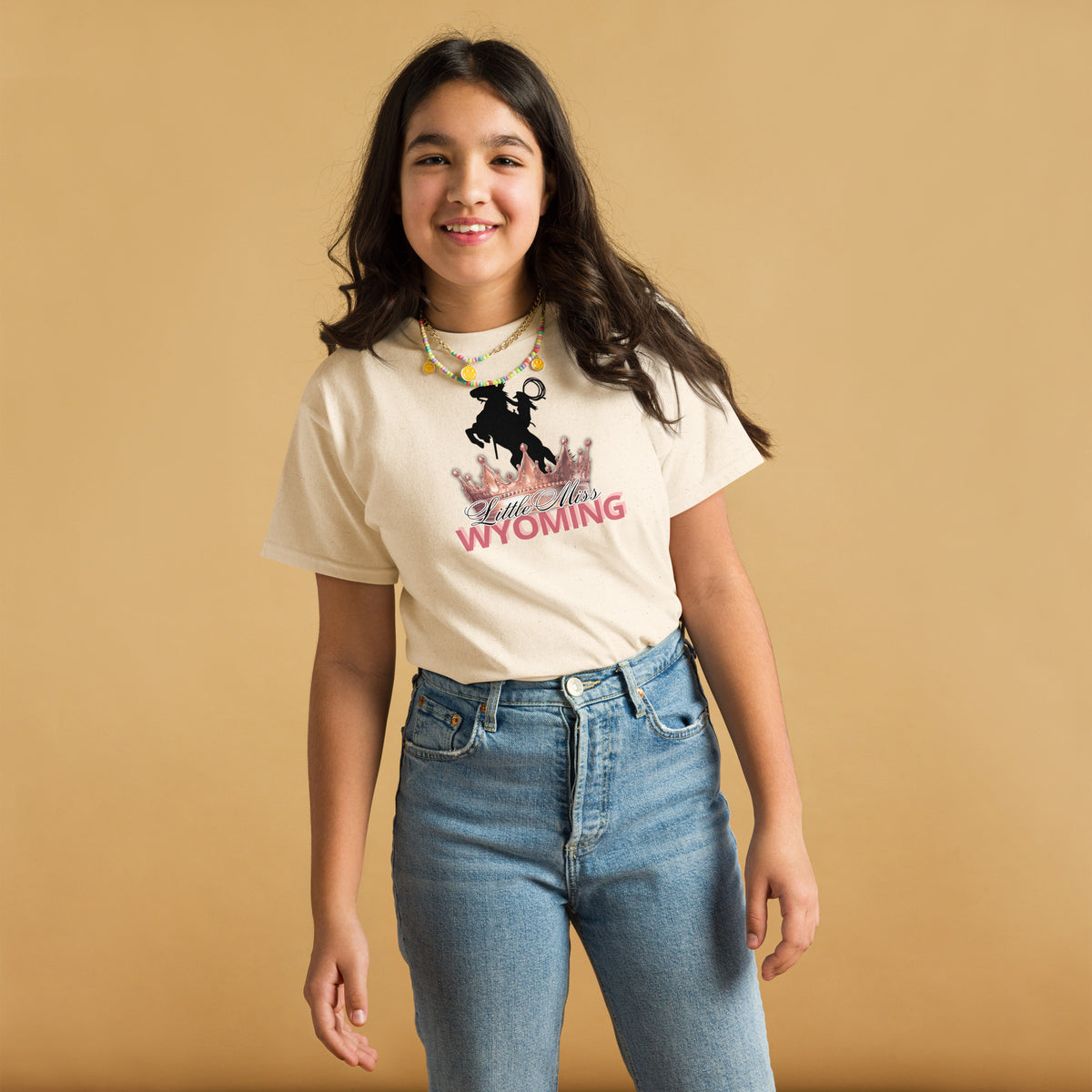 A young girl with long dark hair stands against a solid tan background, wearing a cream-colored graphic t-shirt that says 'Riding High in WYOMING' with a cowboy silhouette, paired with blue jeans.